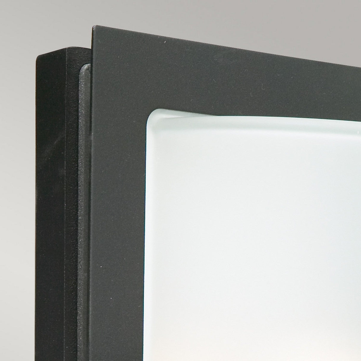 Close-up of a Basel Outdoor 1 Light Wall Lantern in black with frosted glass against a light gray background. The lantern, partially visible, highlights its sleek design and smooth surface. With an IP54 rating, this piece is both stylish and durable.