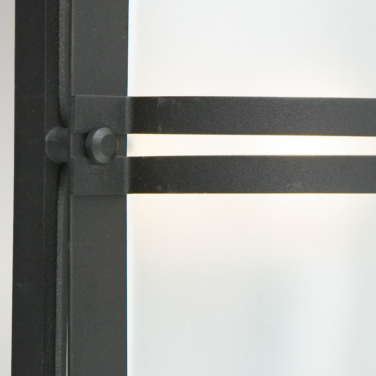 Close-up of the Basel Outdoor 1 Light Wall Lantern in black metal with two horizontal bars, set against a blurred frosted glass background. The lantern features a textured finish and a circular fastener at the intersection.