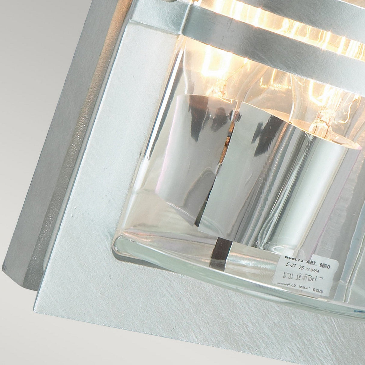 The close-up of the Basel Outdoor 1 Light Wall Lantern - Silver With Clear Glass highlights its modern design. Mounted on a textured wall, the lantern features a sleek silver finish with clear glass and contains a lit bulb that emits a bright light, beautifully illuminating the surroundings.