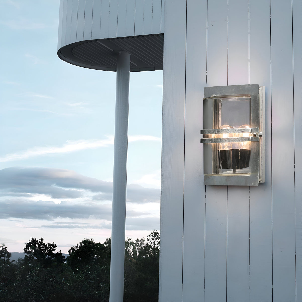 The Basel Outdoor 1 Light Wall Lantern, featuring a silver finish and clear glass, is elegantly affixed to a white wooden wall. As dusk falls, it illuminates the area, highlighting the circular roof structure and silhouetted trees against the cloudy sky.