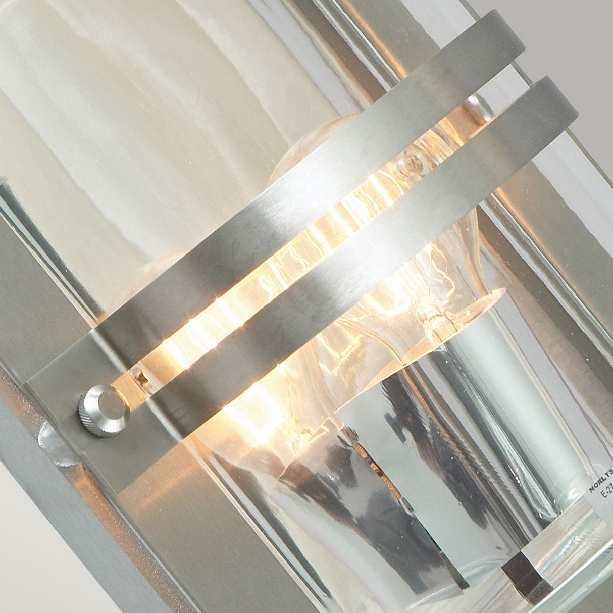 Close-up of the Basel Outdoor 1 Light Wall Lantern, featuring a stainless steel frame and vertical rods encasing a glowing bulb, its light reflecting elegantly off the shiny surface and clear glass design.