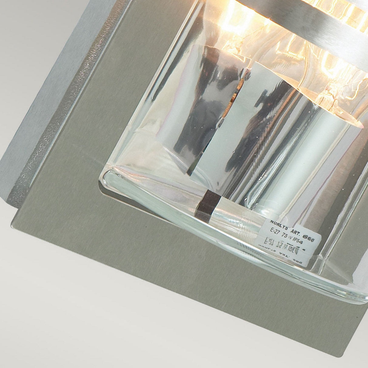 Close-up of the Basel Outdoor 1 Light Wall Lantern, crafted with stainless steel and clear glass. This lighting fixture showcases two exposed light bulbs and features sleek, modern lines with metallic elements for a stylish, contemporary look.