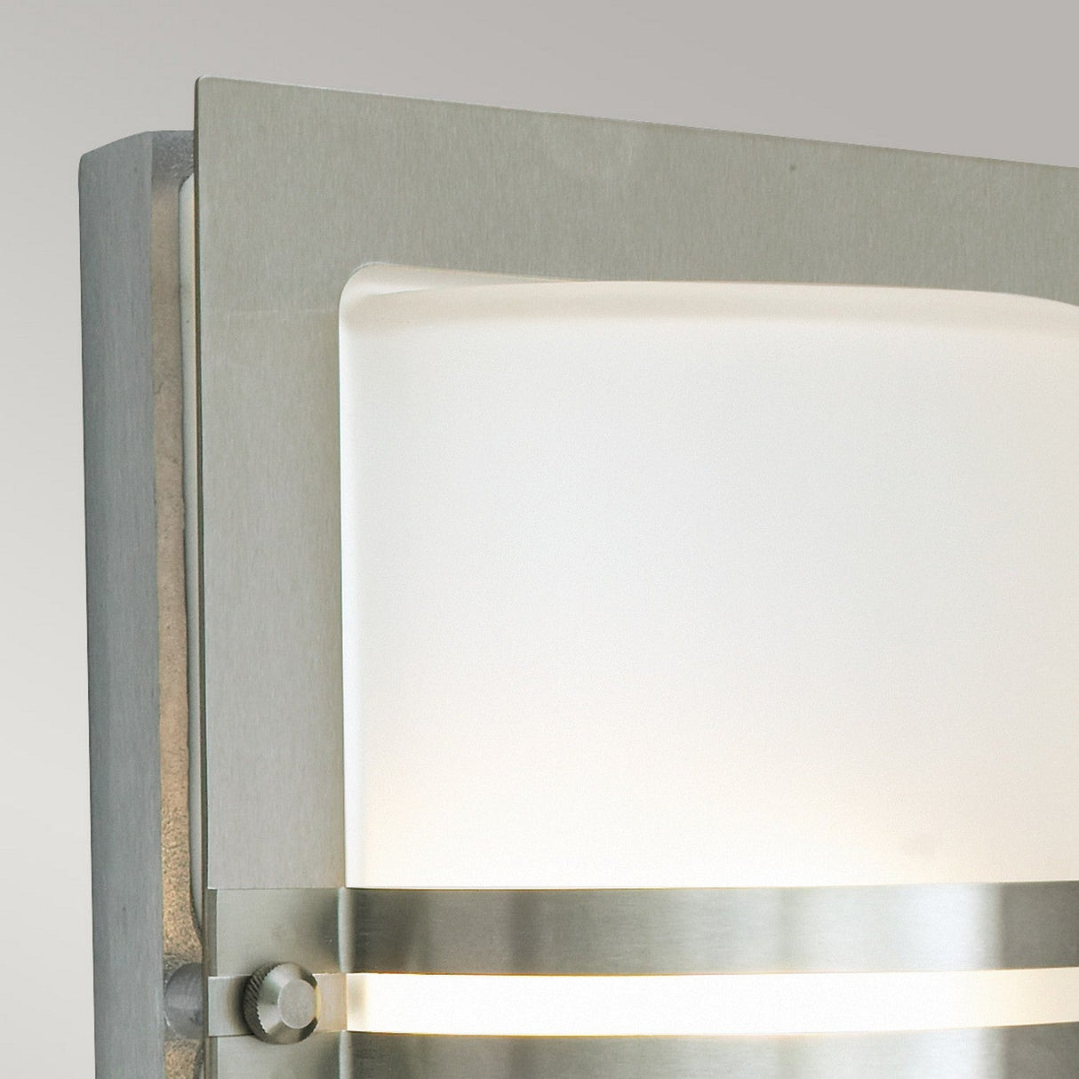 Close-up of a modern wall sconce similar to the Basel Outdoor 1 Light Wall Lantern, featuring a brushed stainless steel frame and frosted glass panel. This design includes two horizontal metal bands and casts a gentle, diffused light against a simple light gray background.