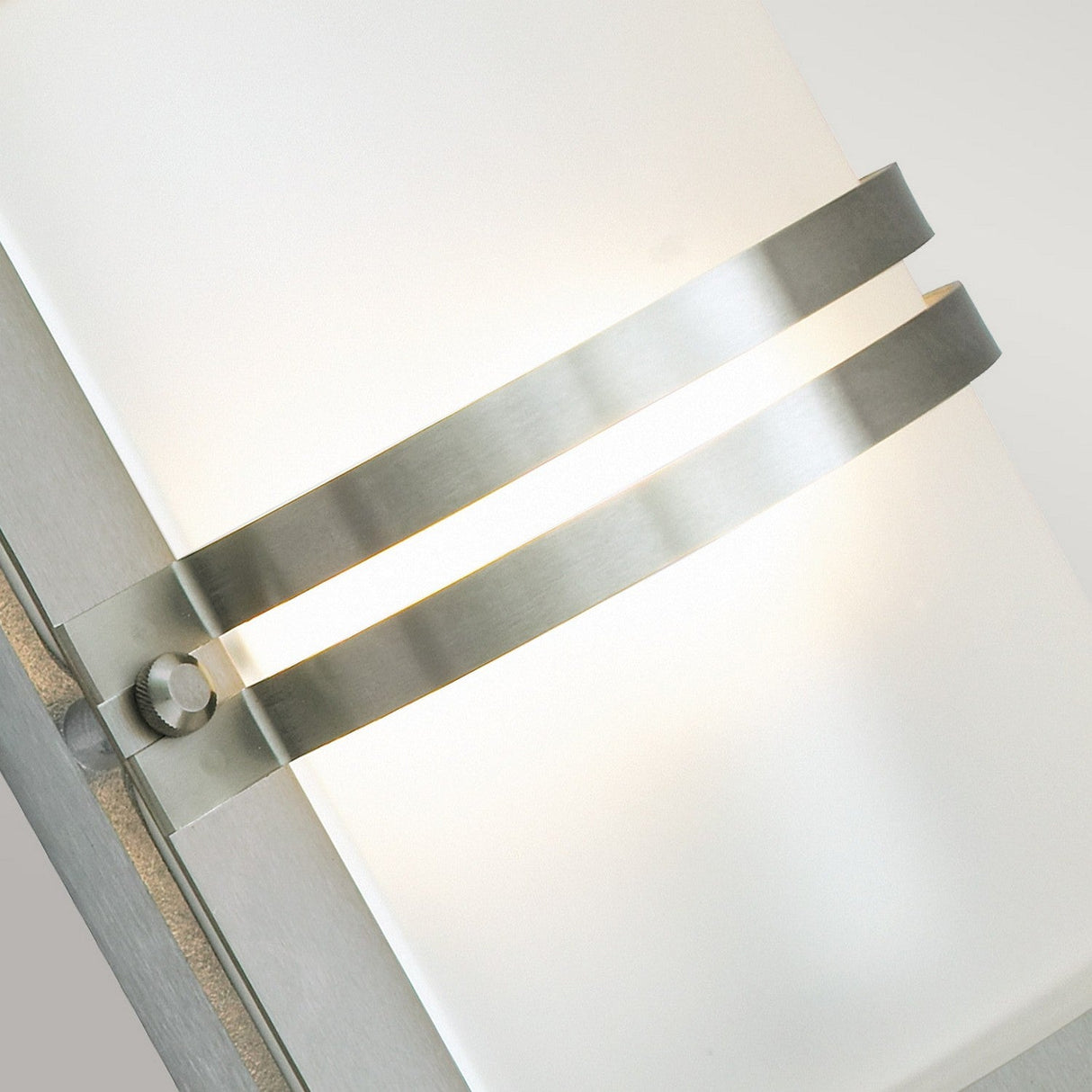 Close-up of the Basel Outdoor 1 Light Wall Lantern - Stainless Steel With Frosted Glass, showcasing its frosted glass panel and stainless steel horizontal bands. The lantern emits a soft glow that highlights its sleek design against a plain background.