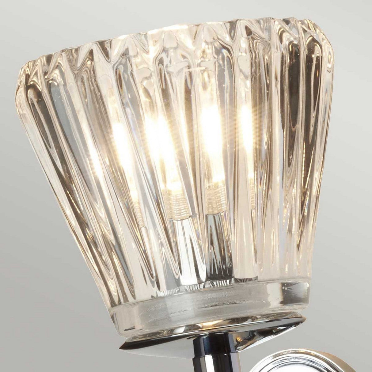 A close-up of the Agatha 2 Light Wall Light - Polished Chrome showcases its modern design with two visible glowing bulbs. The fixture features fluted glass, adding an elegant and contemporary touch, and it is mounted on a sleek polished chrome base with clear cut glass shades.