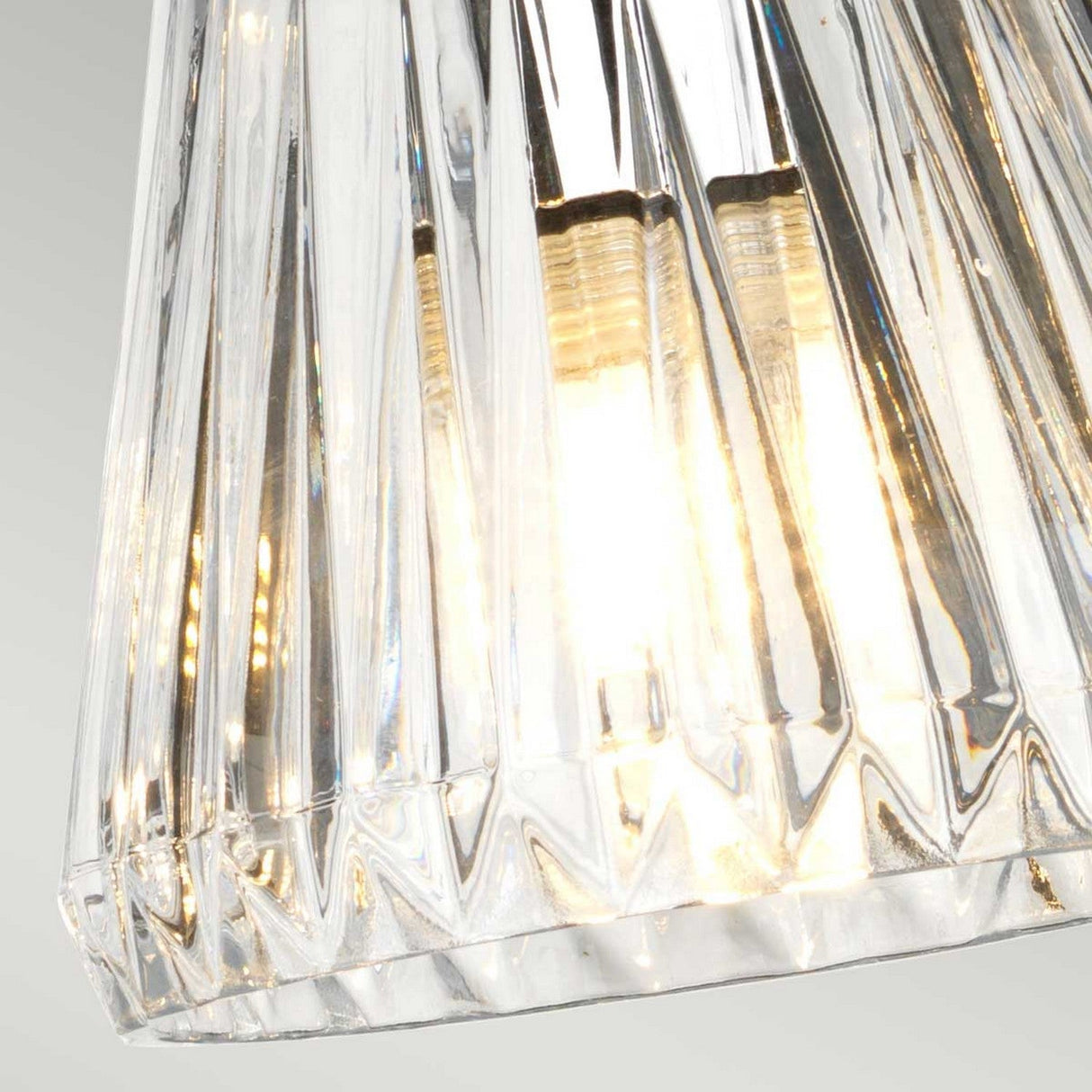 Close-up of the Agatha 3 Light Pendant - Polished Chrome, showcasing a hanging glass light fixture with a faceted design. It is illuminated by the warm glow of the bulb inside. The glass features vertical ridges that shimmer elegantly, enhanced by polished chrome accents for a modern touch.