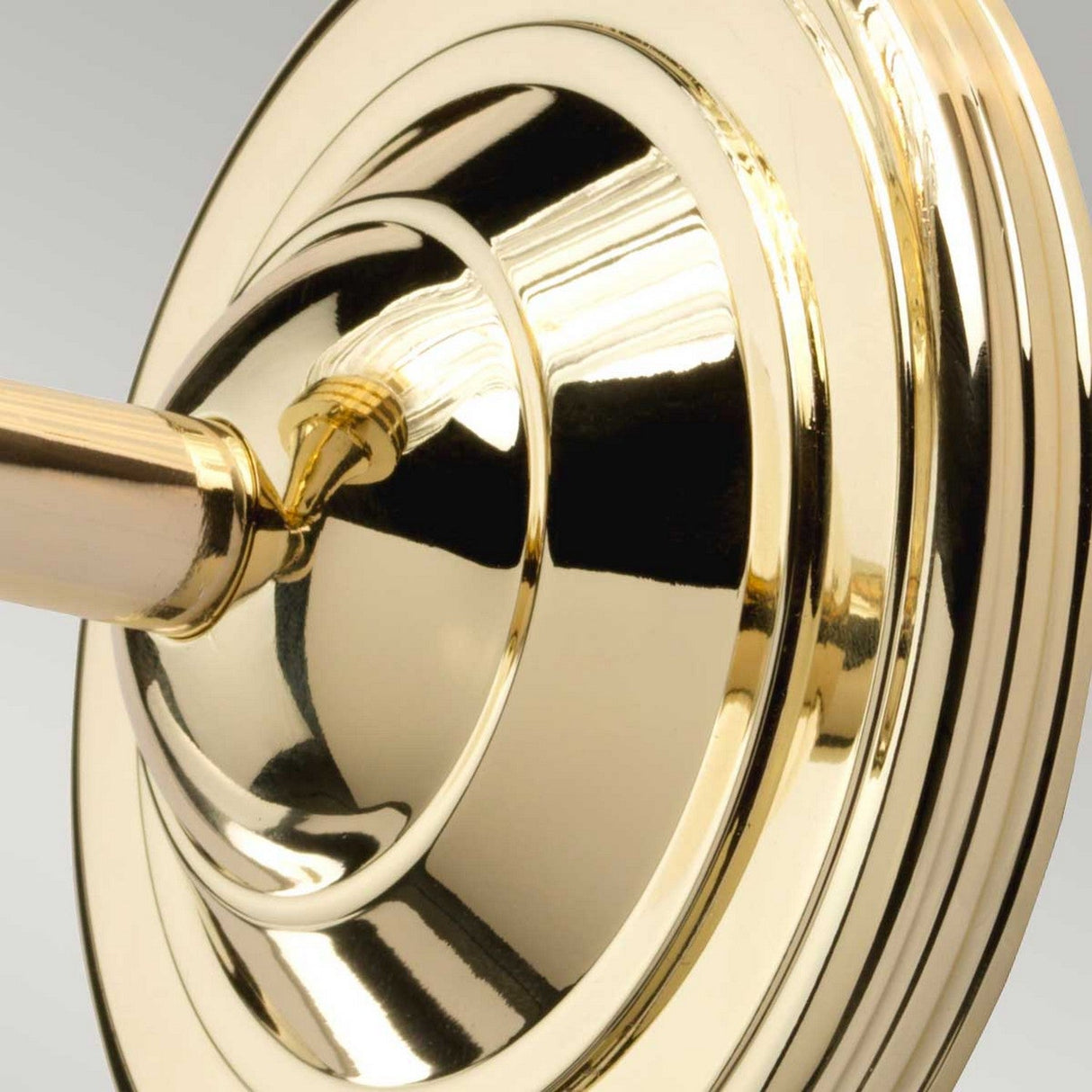 Close-up of an Austen Wall Light in polished brass. The shiny surface reflects light, accentuating its smooth, circular design and intricate details, similar to a polished brass door handle and escutcheon plate. The background features a soft gradient of gray tones.