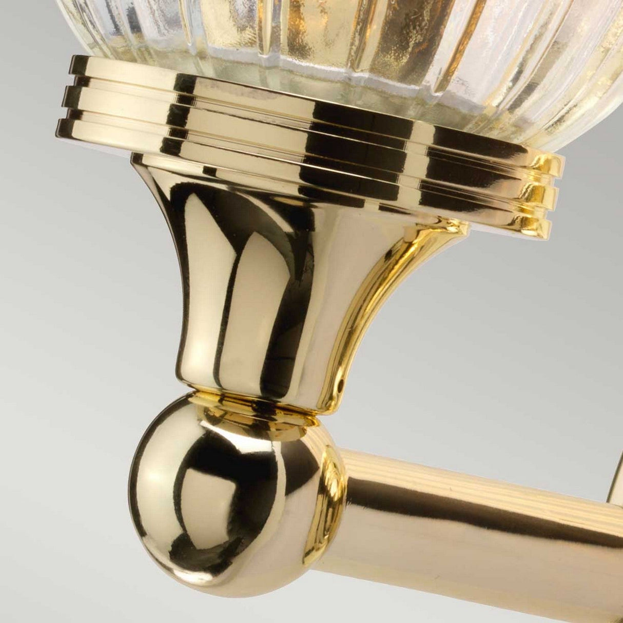 Close-up of the Austen Wall Light - Polished Brass, featuring a spherical base with a grooved, cylindrical upper section seamlessly attached to a ribbed glass component. The image showcases the metallic sheen and detailed craftsmanship characteristic of high-quality wall lights.