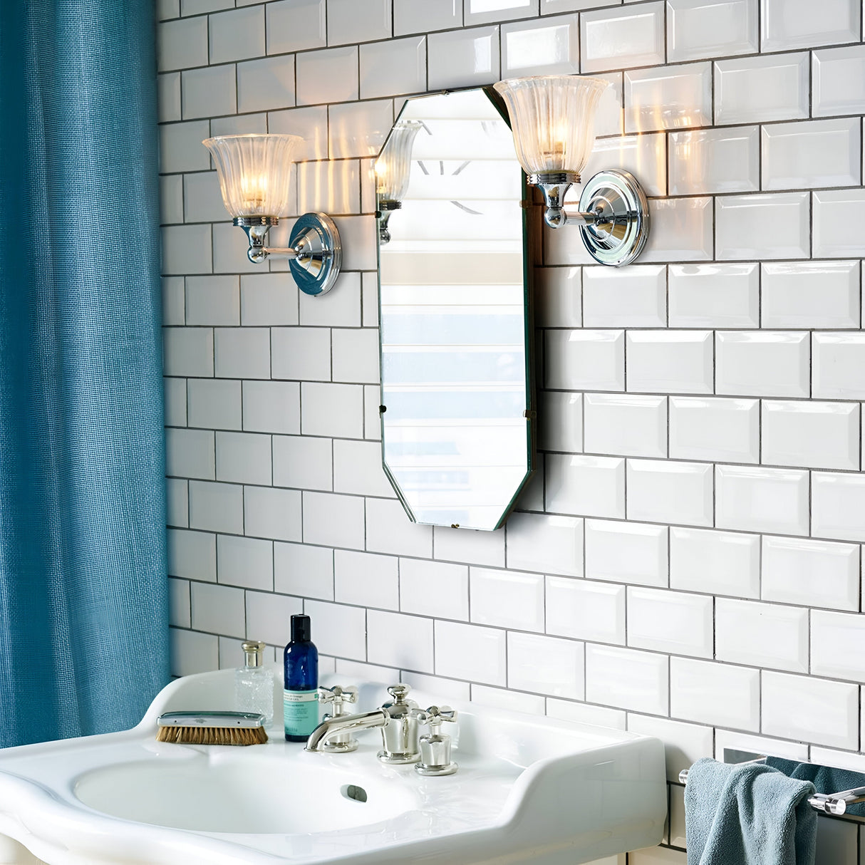A bathroom showcases a wall of white subway tiles, a vintage-style mirror accompanied by twin sconces in polished chrome, and a white ceramic sink outfitted with silver faucets. Toiletries are placed beside an Austen-1 Wall Light - Polished Chrome, while to the left, a blue curtain is partially visible.