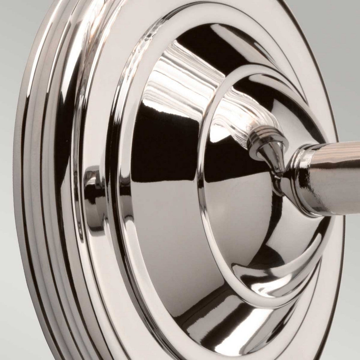 A close-up of a polished nickel wall mount for a showerhead, adorned with concentric circular designs and a brushed texture, beautifully reflects light. This installation is set against a plain background, reminiscent of the sheen characteristic of the Austen Wall Light in Polished Nickel.