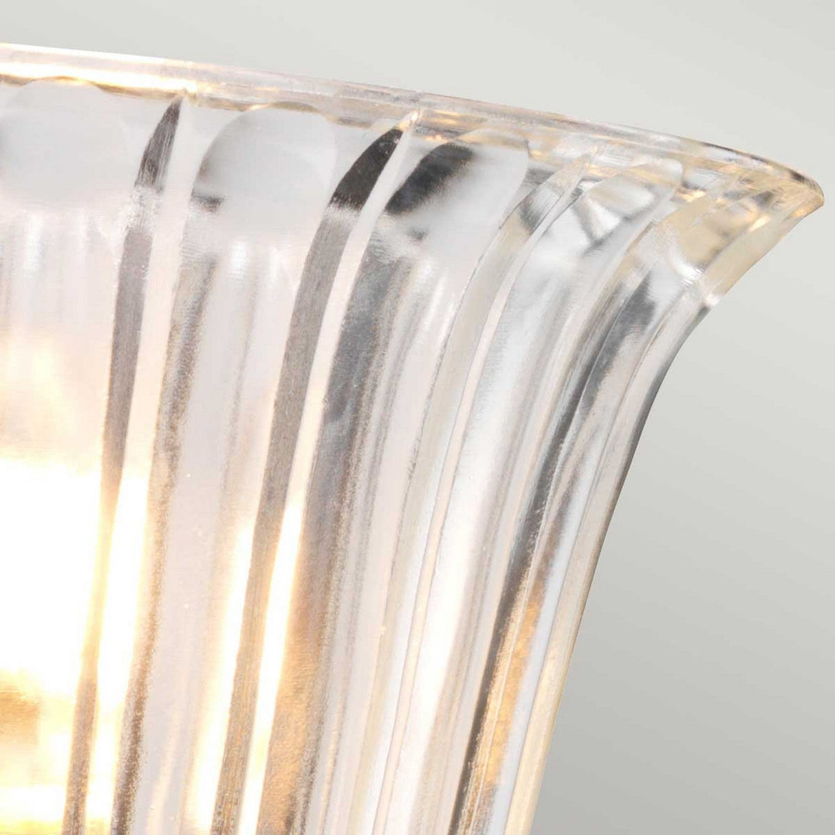 A close-up of the translucent, ribbed glass lampshade, beautifully illuminated from within the Austen Wall Light - Polished Nickel, reveals energy-efficient lighting that casts a warm glow, emphasizing the vertical grooves in the glass. The polished nickel finish brings elegance to a serene and minimalistic setting with a softly blurred background.