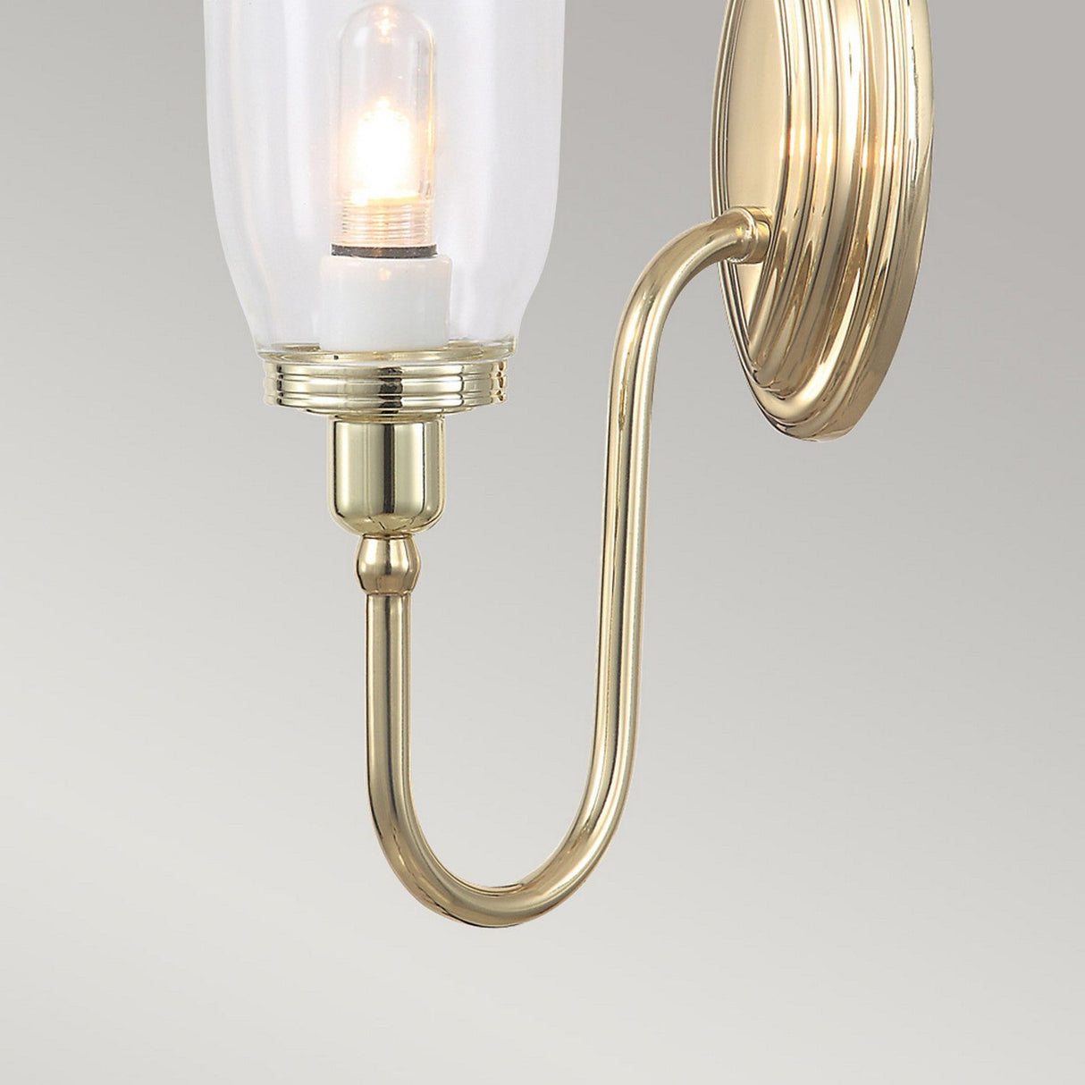 The Blake Wall Light - Polished Brass showcases a polished brass sconce with a gracefully curved arm, which holds a clear glass shade encasing an LED G9 lamp. This sophisticated light fixture is mounted on a round brass base and set against a light gray background.