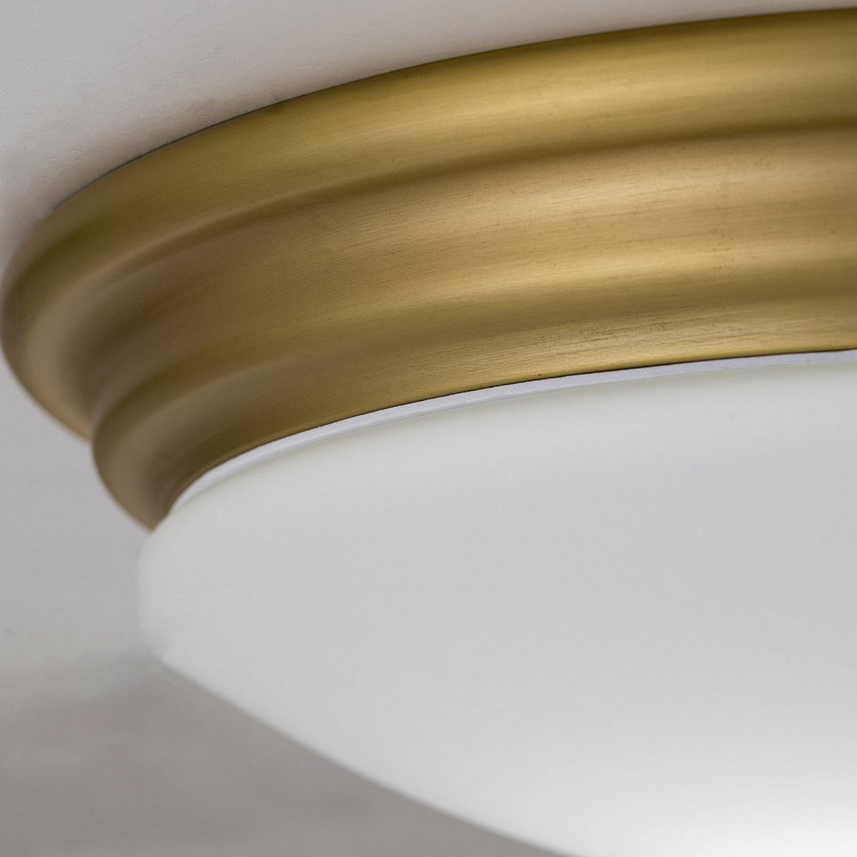 Close-up of the Brompton Flush Light - Brushed Brass, featuring a round brushed brass base paired with a smooth frosted glass dome. This elegant light fixture is mounted on a white ceiling, accentuating its simple yet sophisticated design.