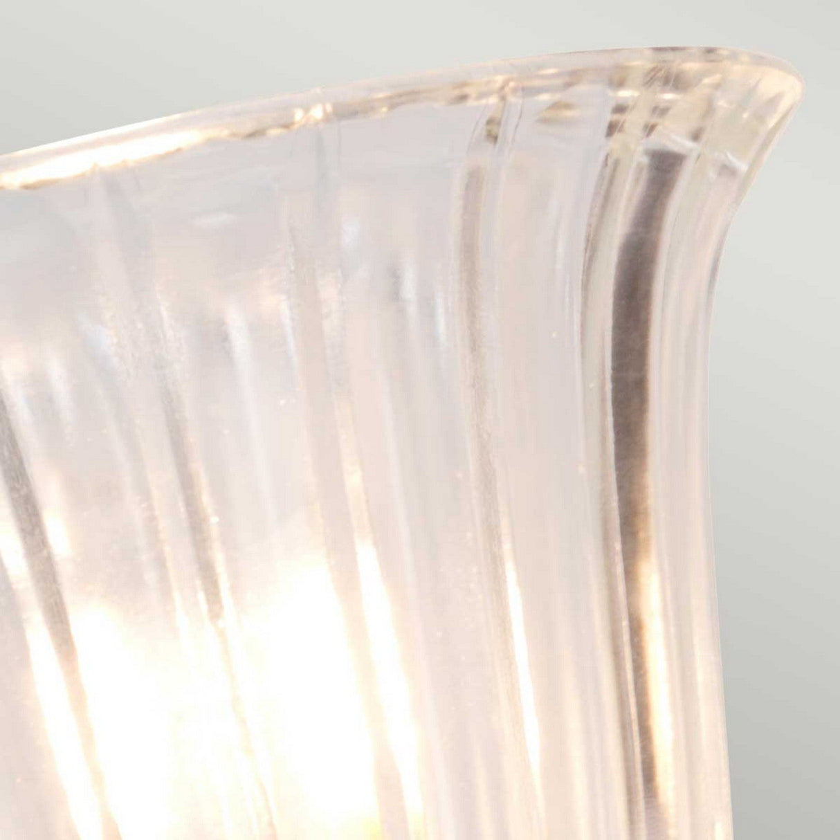 Close-up of a translucent, fluted glass lampshade featuring grooves and a gentle curve, lit from behind by the Carroll Wall Light - Polished Brass, casting a soft glow. This vintage-inspired design brings elegance to the light gray background.