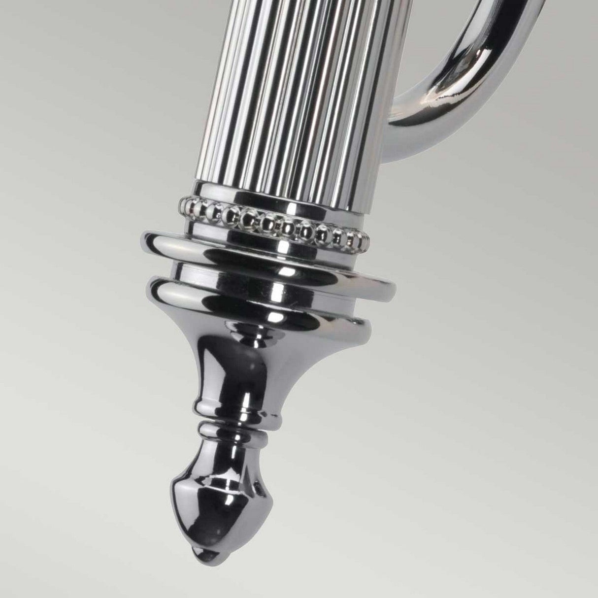 A close-up of the Carroll-1 Wall Light - Polished Chrome showcases an ornate silver metallic piece with a fluted design and decorative beading near the top. The object tapers to a rounded point at the bottom, set against a light gray background.