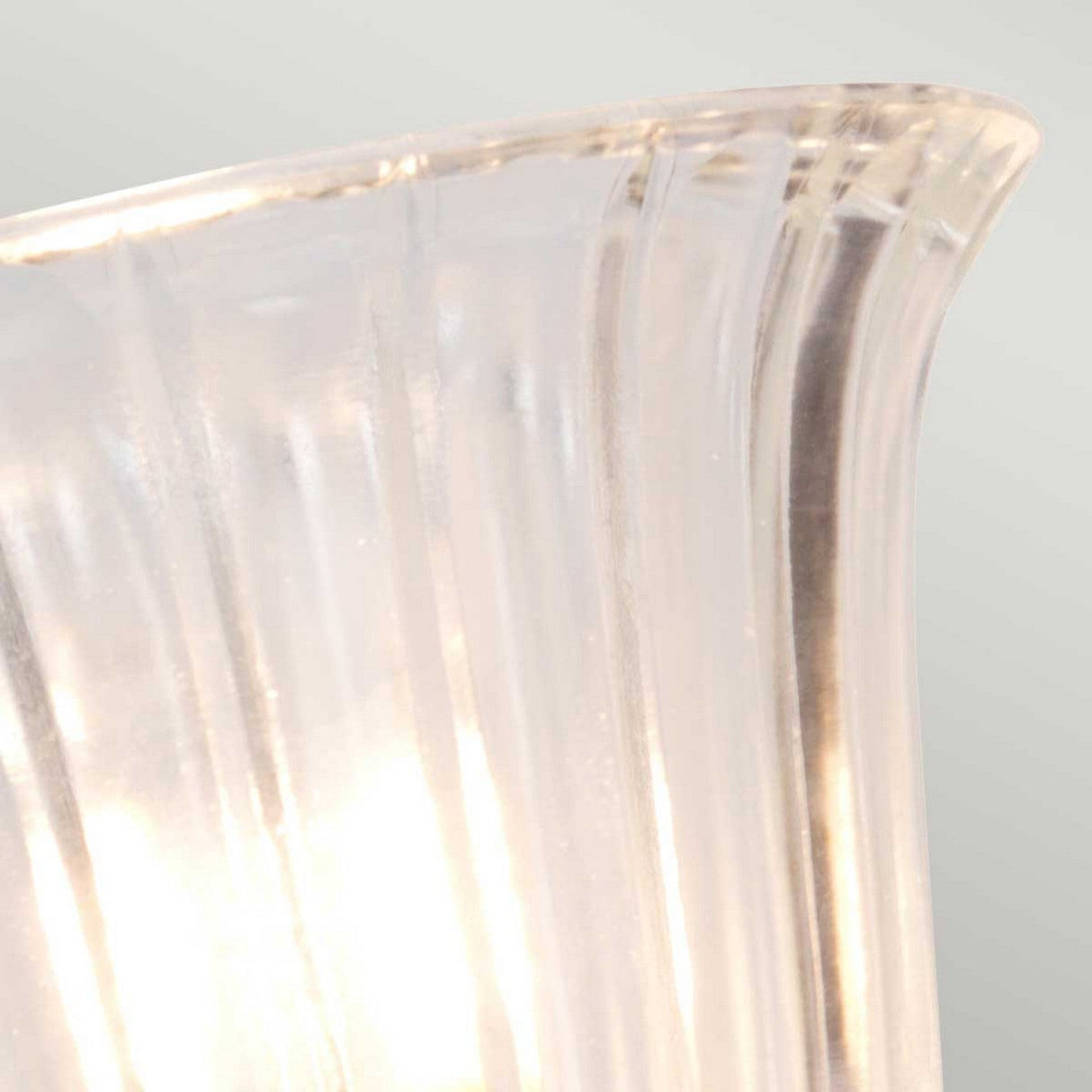 A close-up of the Carroll Wall Light - Polished Nickel reveals a translucent glass lampshade with a vertical ribbed texture, enhancing its polished nickel frame. Soft light filters through, casting a gentle glow against the muted gray background.