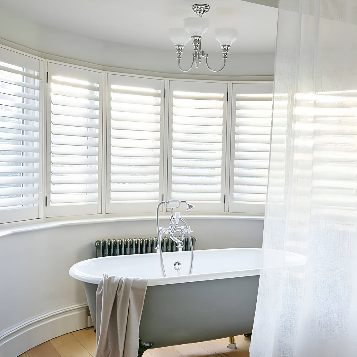 A bright, modern bathroom showcases a freestanding bathtub resting on a wooden floor. Large windows with white shutters pour natural light into the space, complemented by a polished chrome chandelier. The elegance is further highlighted by the Cheadle Wall Light in polished chrome, with its opal glass shade casting an inviting glow while a towel lies gracefully over the tub's edge.