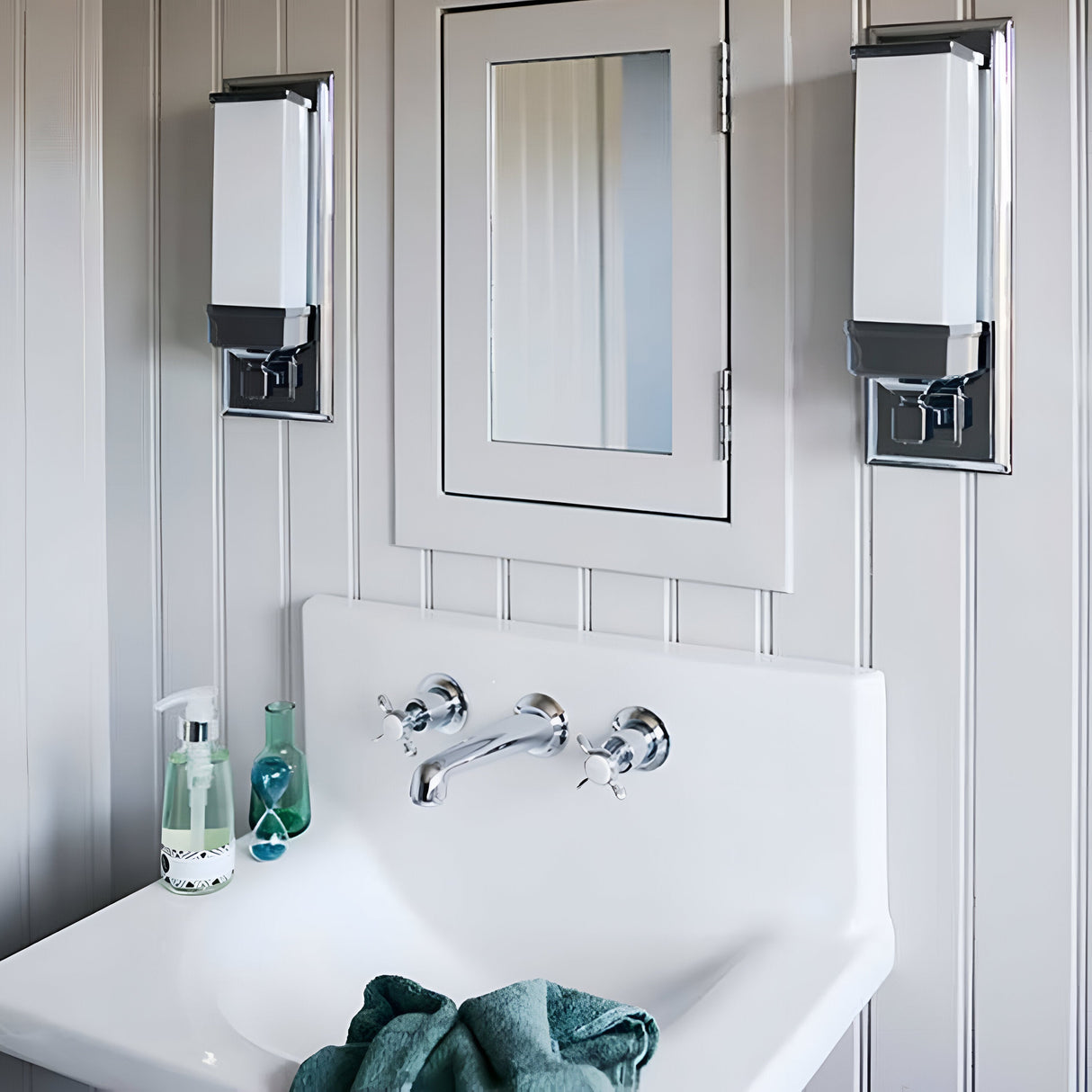 A white bathroom sink with polished chrome faucets is set against a paneled wall, complemented by a mirror cabinet above the sink, flanked by two modern soap dispensers. The blue towel and bottles of soap on the sink are illuminated by Cambridge Wall Lights in polished chrome, each featuring opal glass shades.