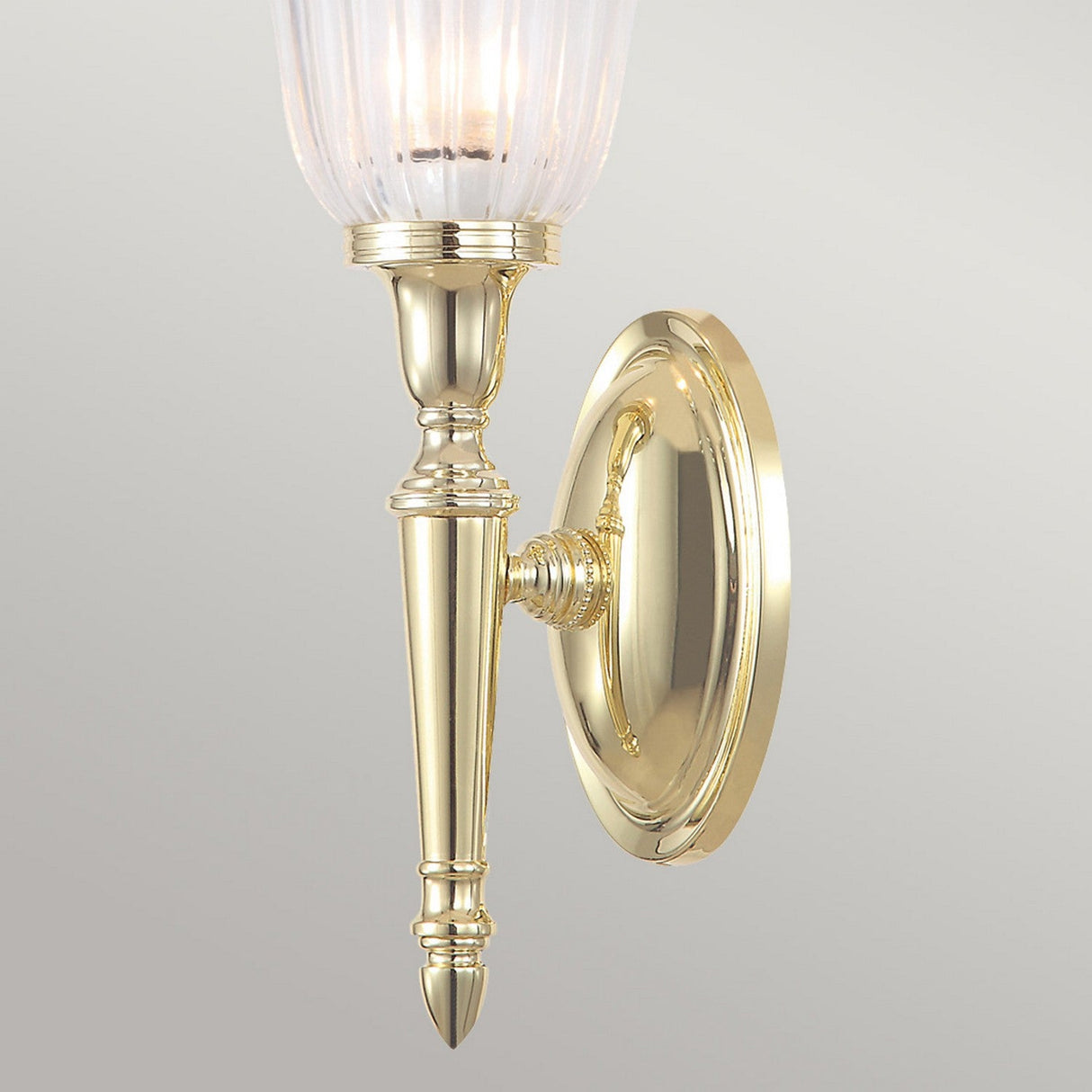 Introducing the Dryden-1 Wall Light in polished brass, characterized by its oval base and sleek vertical design. It boasts a ribbed glass lampshade at the top, using an LED G9 lamp to enhance its classic charm against a neutral gray background.