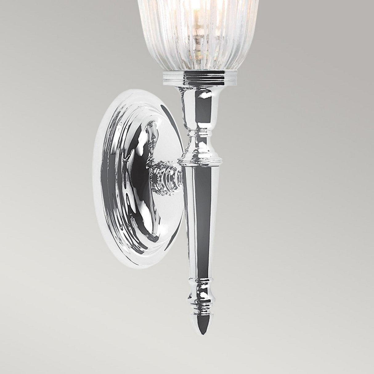 The Dryden-1 Wall Light, featuring a polished chrome finish, is a silver wall-mounted sconce with an elegant tapered design and a fluted glass shade. Its LED G9 lamp softly illuminates the space, with light gracefully reflecting off the surface against a plain gray background.