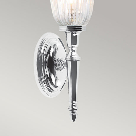 The Dryden-1 Wall Light, featuring a polished chrome finish, is a silver wall-mounted sconce with an elegant tapered design and a fluted glass shade. Its LED G9 lamp softly illuminates the space, with light gracefully reflecting off the surface against a plain gray background.