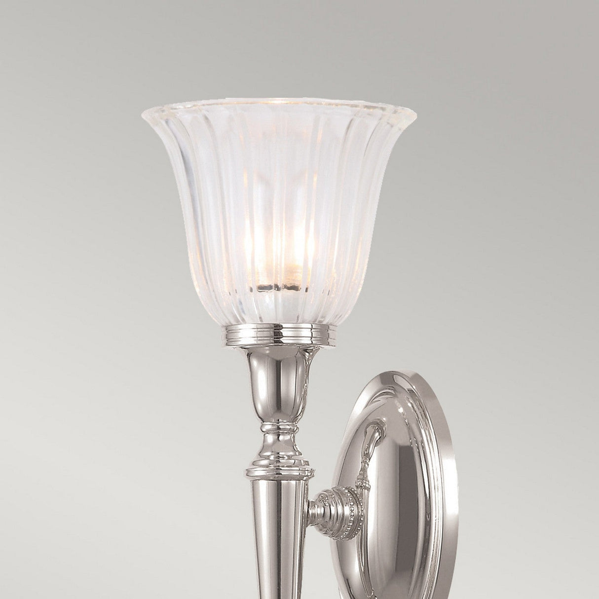 The Dryden-1 Wall Light in polished nickel is a wall-mounted sconce featuring an upward-facing, fluted glass shade that softly illuminates a neutral gray background. It comes with an LED G9 lamp and has an IP44 rating for enhanced durability.