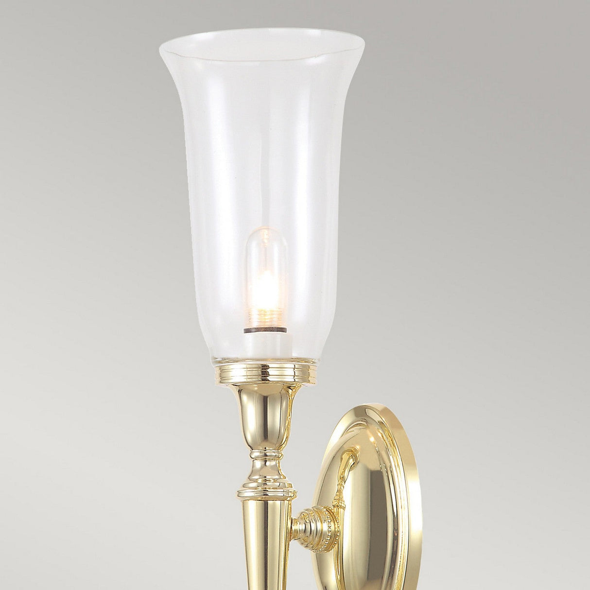 The Dryden-2 Wall Light in Polished Brass showcases a tall, clear glass shade that encases a lit bulb. It is mounted on an oval brass backplate with a polished finish against a neutral background, capturing the charm of traditional interior lighting.