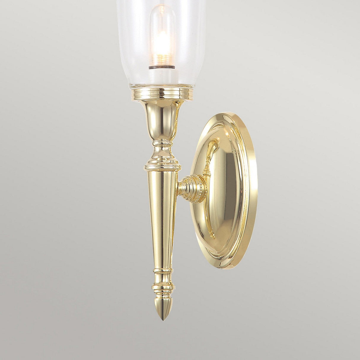 The Dryden-2 Wall Light in Polished Brass showcases a tall, clear glass shade with a visible bulb, elegantly designed for traditional interiors. It features intricate detailing and a polished finish, all mounted on a round brass base.