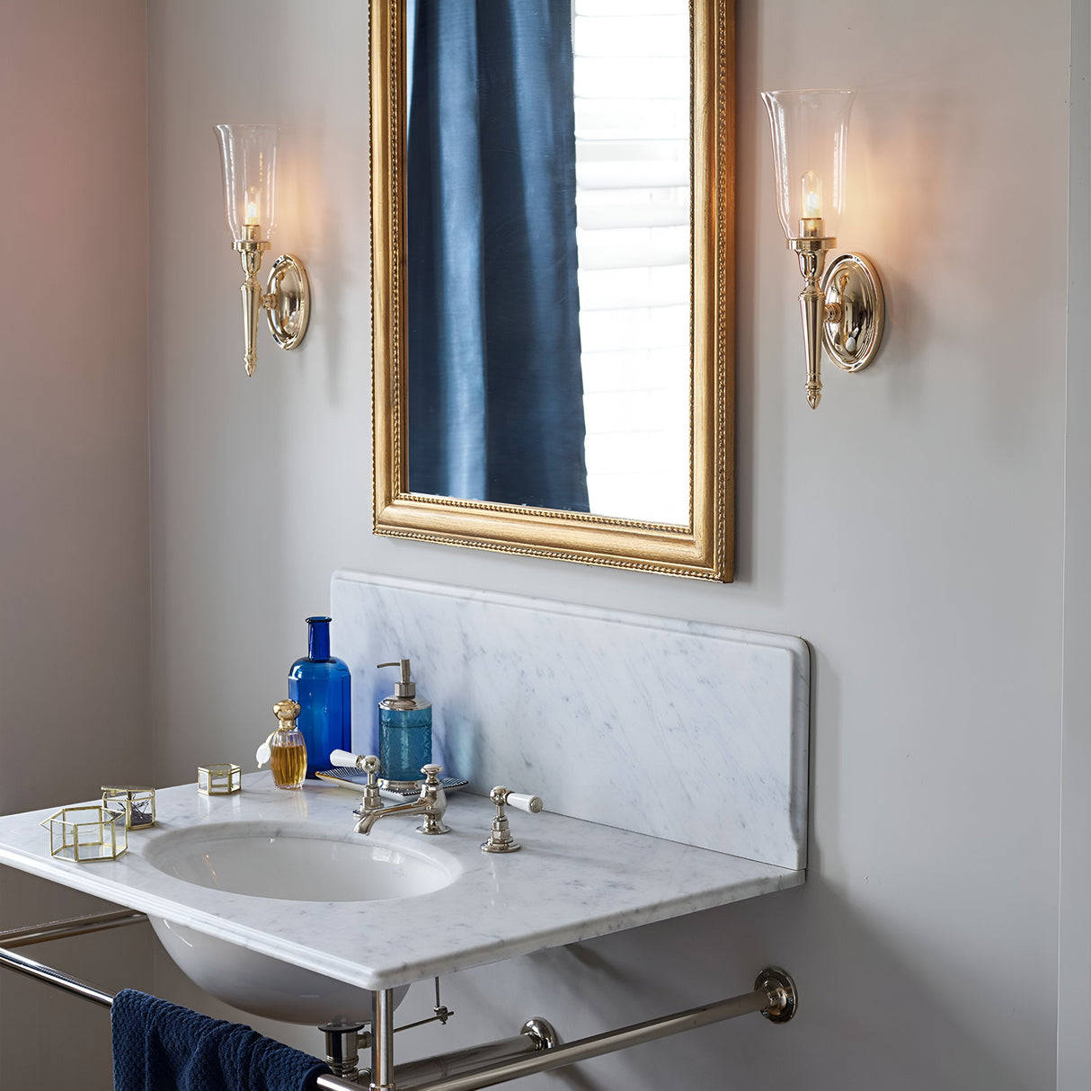 The bathroom showcases a marble countertop and sink, complemented by two Dryden-2 Wall Lights in polished brass with glass covers flanking a framed mirror. The countertop is adorned with blue and gold decor items, while a blue curtain adds an elegant touch in the background.