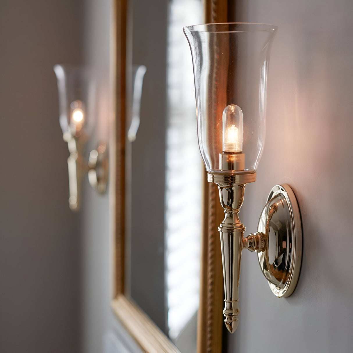 The Dryden-2 Wall Light in polished brass, featuring a glass shade, illuminates the space and reflects beautifully in a framed mirror, enhancing the depth of the room. Its energy-efficient LED lamps provide a soft glow that creates a warm and inviting ambiance.