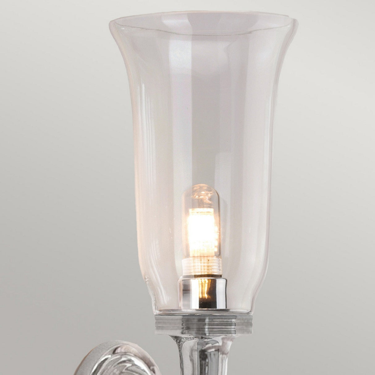 A close-up of a lit LED G9 lamp inside a transparent, tulip-shaped glass lamp shade showcases a sleek and modern design with a frosted base and polished chrome accents, reminiscent of the elegant Dryden-2 Wall Light - Polished Chrome, set against a plain light gray background.