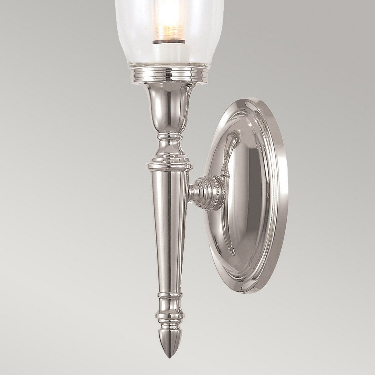 The Dryden-2 Wall Light in polished nickel boasts a classic design with an upward-facing glass shade and is mounted on a circular backplate. Its LED G9 lamp is illuminated, providing a warm glow.