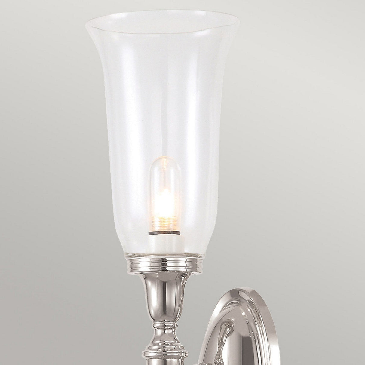 The Dryden-2 Wall Light in Polished Nickel is a stylish wall-mounted fixture featuring a tall, translucent glass shade. It is illuminated by LED G9 lamps that cast a warm glow against the plain, light-colored wall.