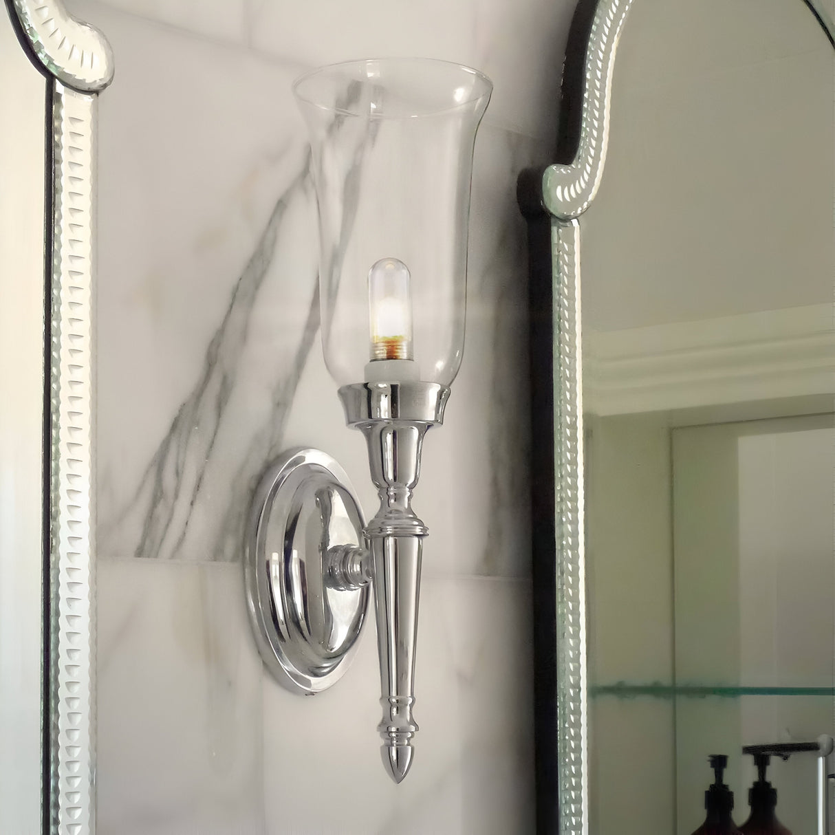The Dryden-2 Wall Light in polished nickel, featuring a clear glass lamp shade, is affixed next to a decorative mirror. The background showcases a marble-patterned wall, creating a classic and elegant ambiance highlighted by LED G9 lamps.