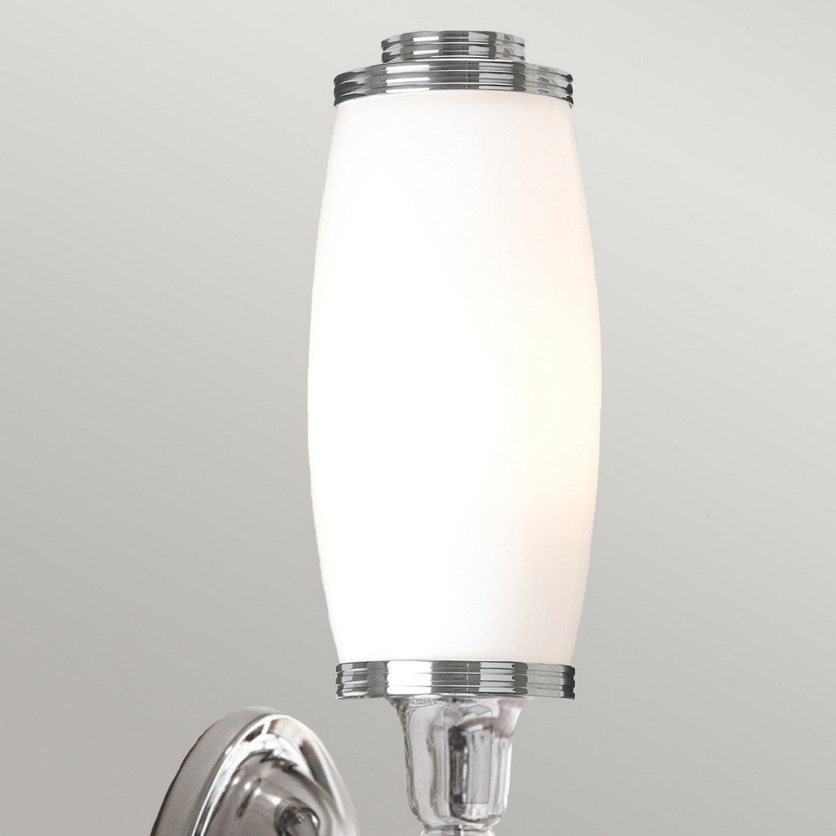 Featuring the Dryden-4 Wall Light in polished chrome, this setup includes an elongated white glass shade that stands out against a light gray wall, showcasing its sleek and modern design.