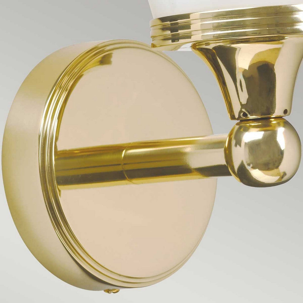 Close-up of the Eliot 1 Light Wall Light in polished brass, perfect for bathroom lighting, showcasing a round base. It features a gleaming surface with a cylindrical arm that extends to a decorative spherical joint supporting an LED fixture, partially obscured in the image.