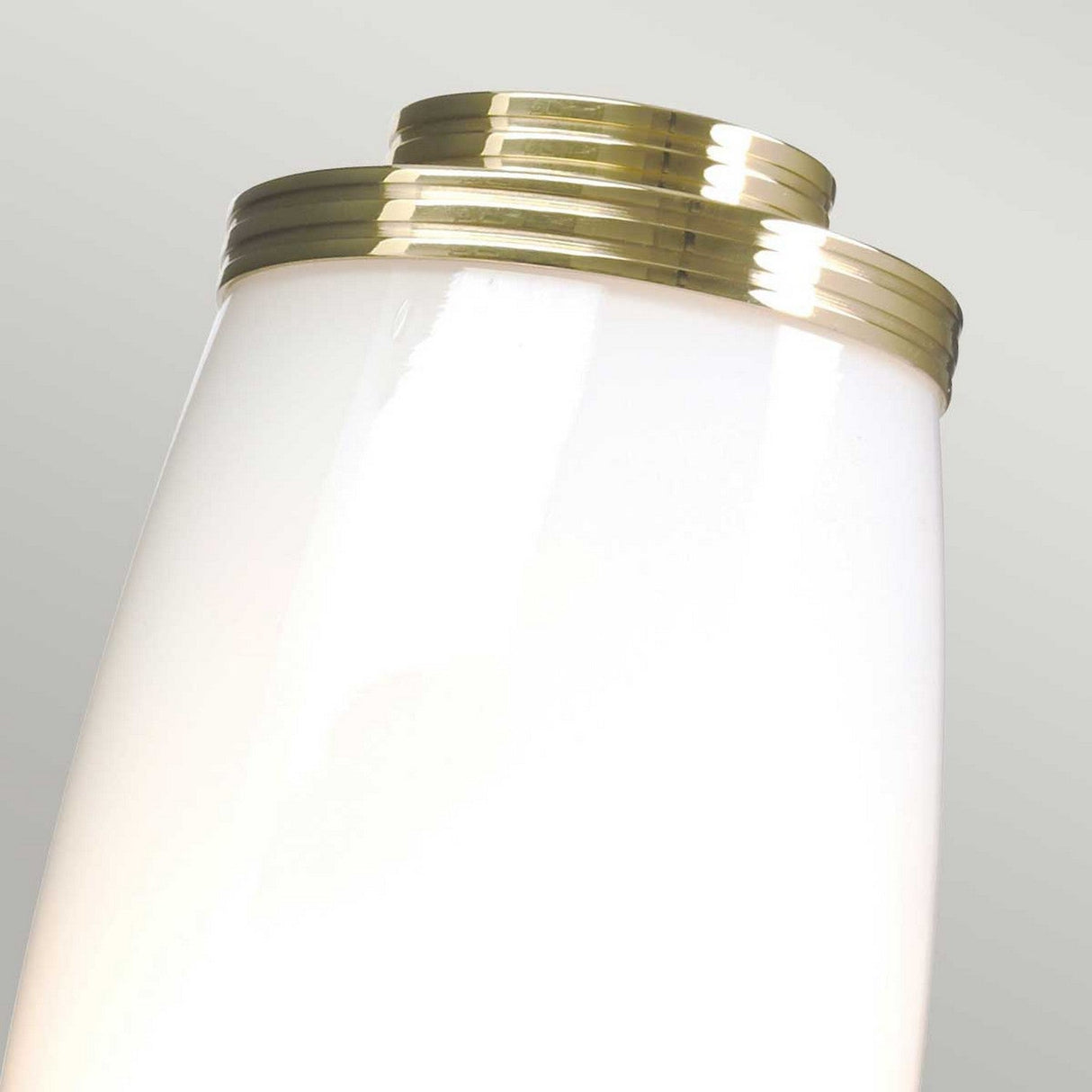 Close-up of a contemporary white glass vase featuring gold accent rings near the top, beautifully illuminated by the Eliot 1 Light Wall Light in polished brass, set against a light gray background.