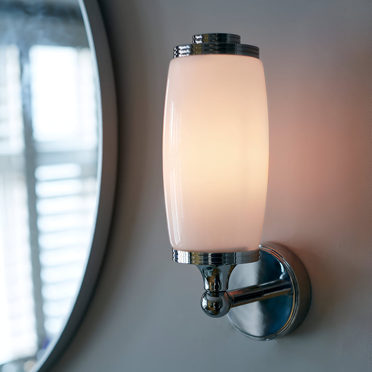 The Eliot 1 Light Wall Light - Polished Chrome, featuring a smooth, cylindrical white shade and polished chrome accents, is illuminated. It is installed next to a large, round mirror reflecting window blinds in a softly lit room, making it an ideal choice for bathroom lighting with its efficient LED G9 lamp.