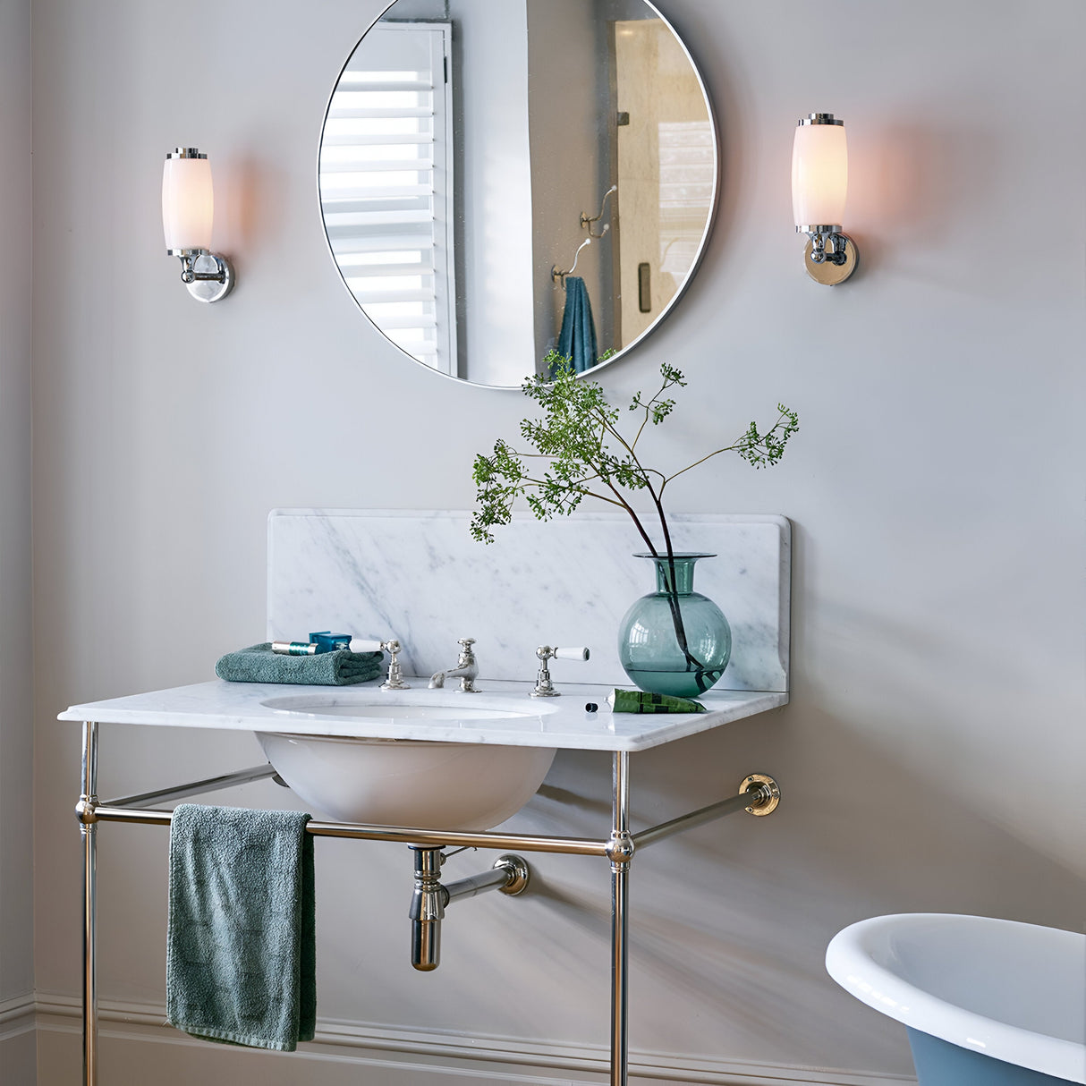 In a contemporary bathroom setting, a round mirror is positioned above a marble sink with metallic legs. Two Eliot 1 Light Wall Lights in Polished Chrome frame the mirror, enhancing the bathroom's illumination. A green vase filled with branches rests on the counter, alongside a towel draped over the sink, while part of a bathtub can be seen in the foreground.