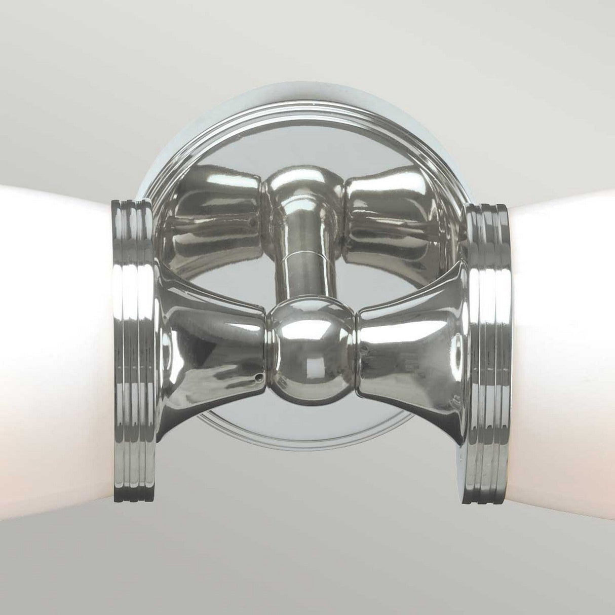 Close-up of the Eliot 2 Light Wall Light in polished chrome, featuring energy-saving LED lamps within two white cylindrical designs extending on either side. The fixture has a circular chrome base and symmetrical design, offering a modern and elegant look against a light gray background, making it ideal for bathroom lighting.