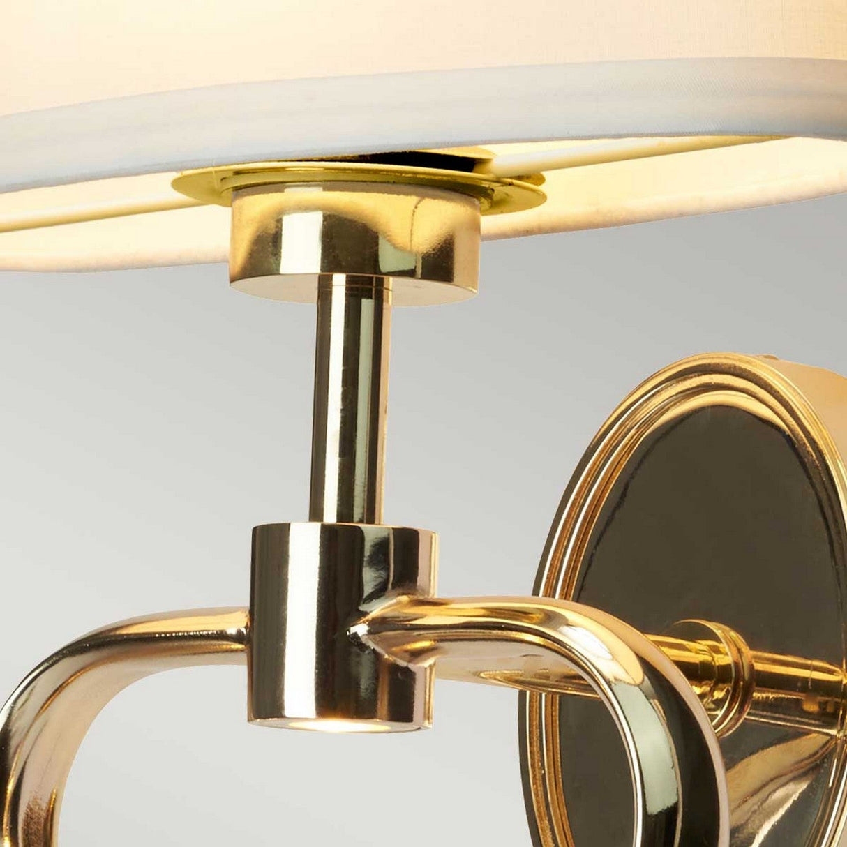 Close-up of the Falmouth Wall Light - Gold/White Shade. The fixture boasts polished gold curved arms and a soft white lampshade, establishing a warm and sophisticated ambiance against a neutral gray background.