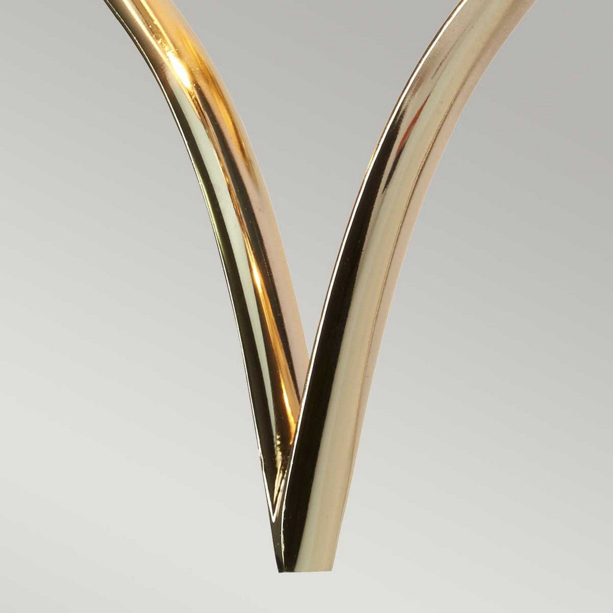 Close-up of the Falmouth Wall Light - Gold/White Shade featuring two metallic, golden curved rods with a gold finish creating a symmetrical V shape that resembles a heart-shaped frame against a light gray background.