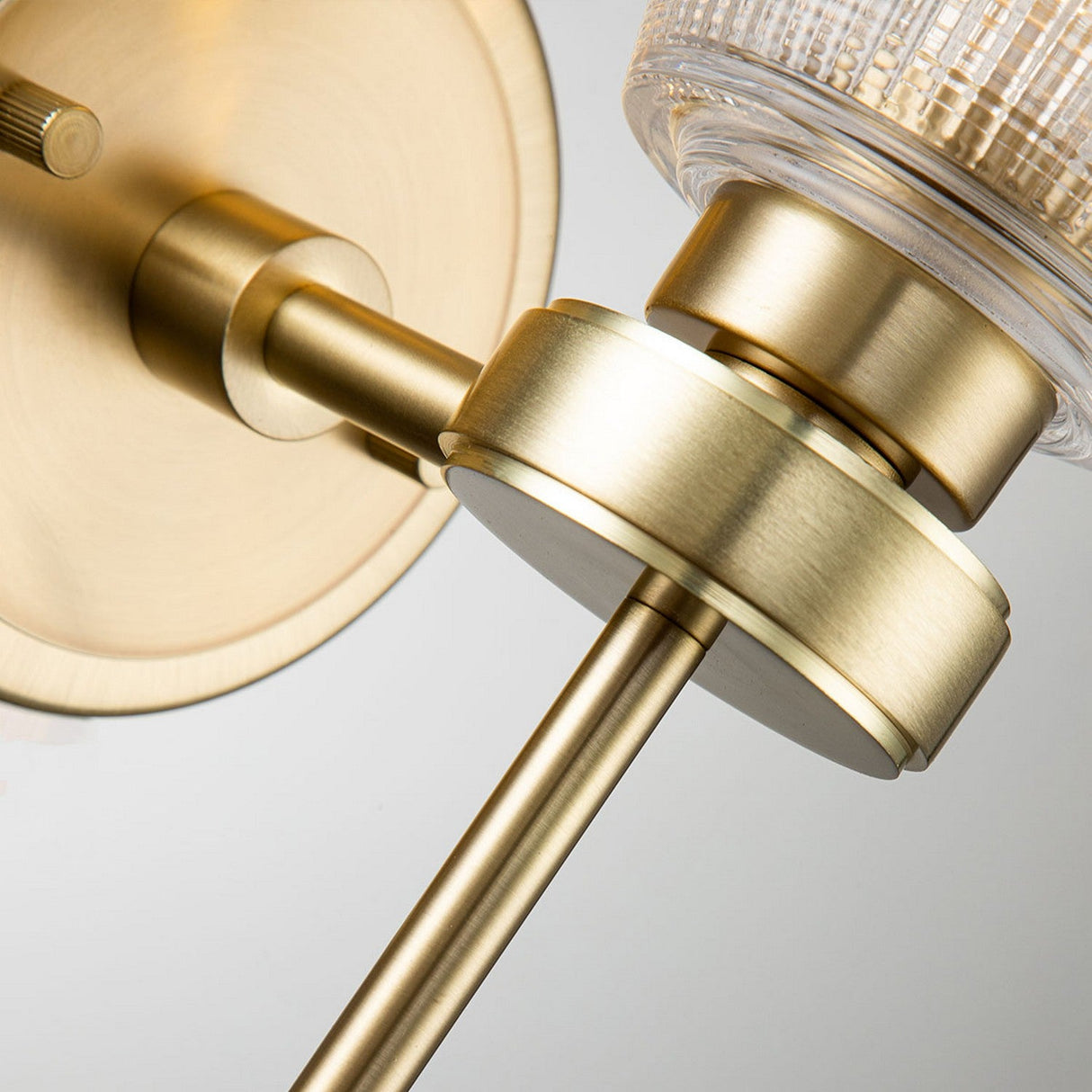 A detailed view of the Gunnislake 1 Light Wall Light in brushed brass highlights its sleek, cylindrical and round elements, complemented by a prismatic glass shade. This modern and minimalistic design exemplifies Elstead Lighting's craftsmanship.