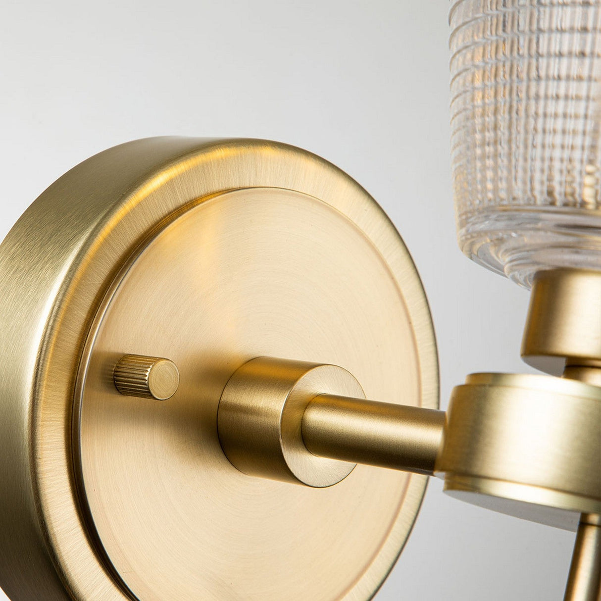 Close-up of the Gunnislake 1 Light Wall Light in brushed brass by Elstead Lighting, featuring a modern sconce with a prismatic glass shade. The design showcases a cylindrical arm extending from the round base, emphasizing its sleek and elegant appeal.