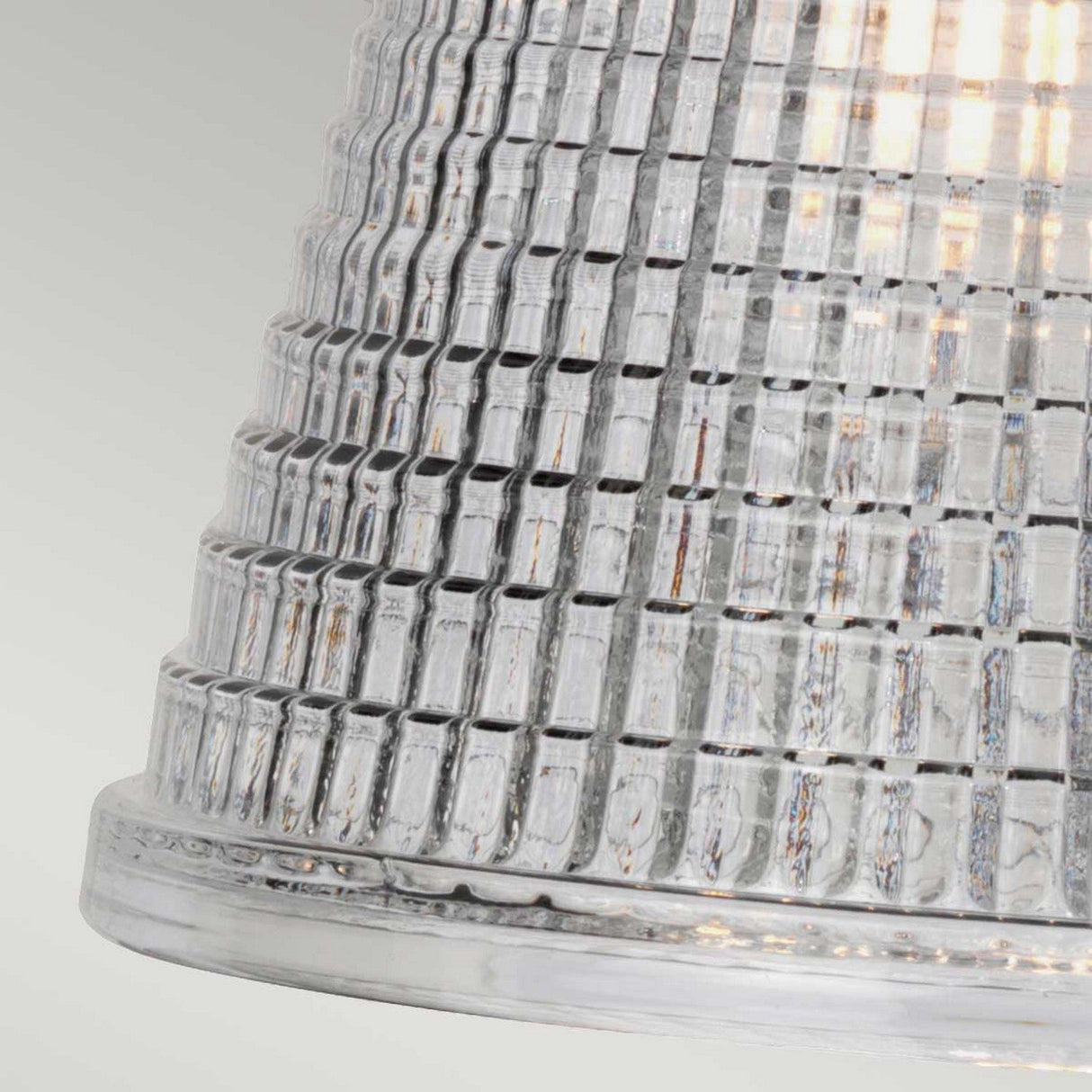 A close-up of the Gunnislake 1 Light Wall Light in polished chrome reveals its textured, prismatic glass lamp shade, glowing with a soft, warm illumination. The design features vertical ridges and horizontal grooves that form a grid-like pattern, set against a backdrop of soft pale green that highlights its polished chrome accents.