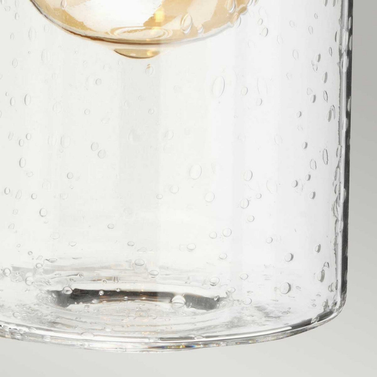 Close-up of a clear glass with a textured, bubbly surface. The background features a soft gradient, accentuating the vintage-inspired lighting effect. Inside, there's a partial view of an amber-colored object resembling the Morvah 1 Light Outdoor Wall Light in brushed brass finish.