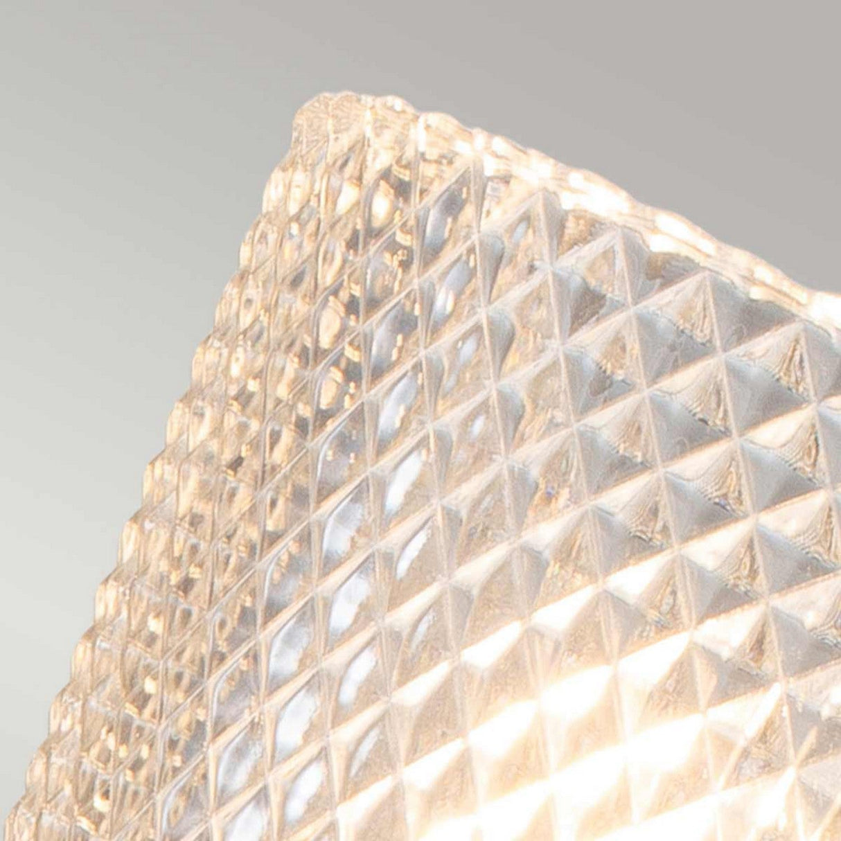 A close-up of a textured, translucent glass surface with a diamond pattern is beautifully illuminated by the Verity 1 Light Wall Light - Brushed Brass. This energy-efficient LED lamp casts a warm, diffused glow that accentuates the intricate details of the glass design.