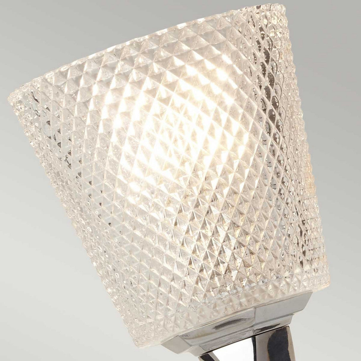 A close-up of the lit Verity 1 Light Wall Light in polished chrome, featuring a textured, diamond-patterned glass shade in modern interiors. The energy-efficient illumination is bright, emphasizing the intricate design of the glass, while the background is softly blurred in gray tones.