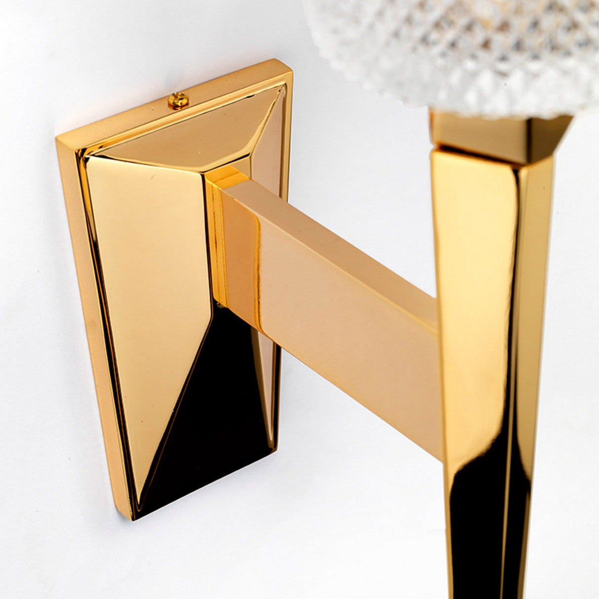 Close-up of the Verity 1 Light Wall Light in polished gold, showcasing a modern, gold-colored metal sconce with an angular, geometric design. The glossy finish of the fixture is mounted against a light background, featuring an energy-saving LED lamp that highlights the textured glass element at the top.