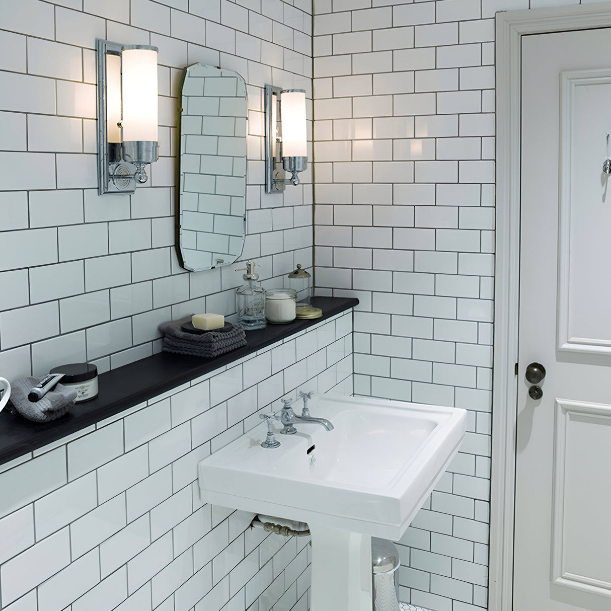 A modern bathroom with transitional design showcases white subway tiles, a rectangular pedestal sink, and a small oval mirror. Flanking the mirror are two Worcester 1 Light Wall Lights in polished chrome finish. Above the sink, a shelf holds toiletries and towels, while a white door is positioned on the right.